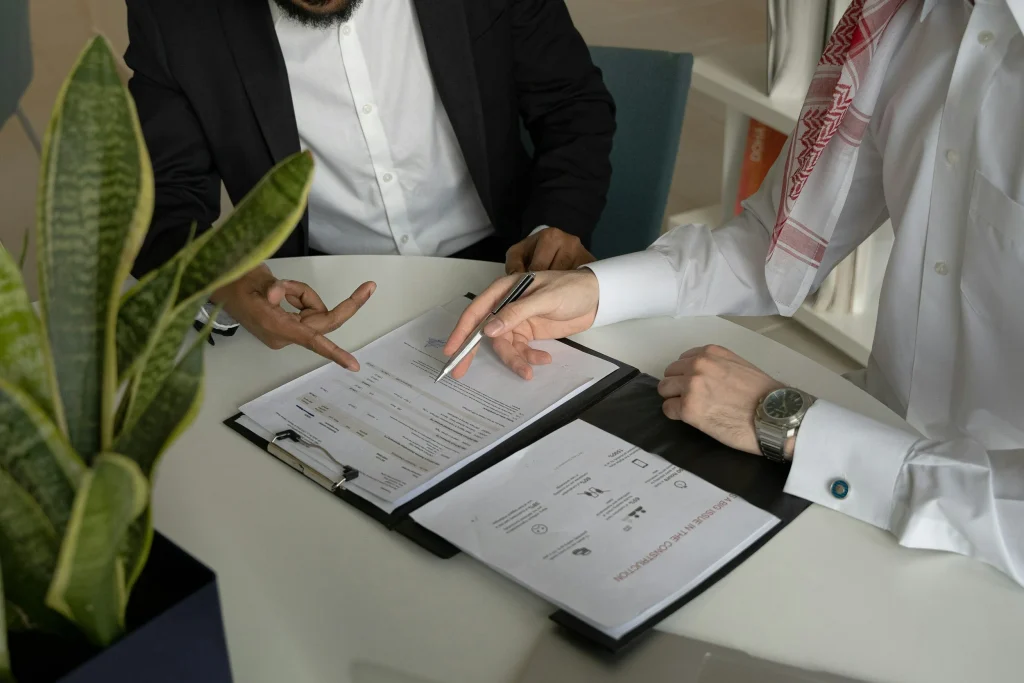 Two men in business attire discussing documents at a table in an office, one pointing at a paper and the other holding a pen, with a potted plant in the foreground.