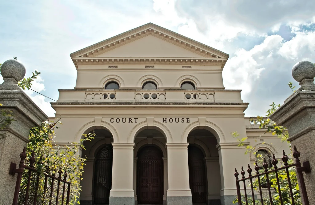 A historic courthouse with white walls, arched doorways, and a triangular pediment, viewed from the front with a fence and greenery in the foreground, under a partly cloudy sky.